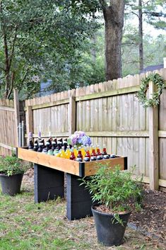 an outdoor table with bottles and flowers on it in front of a wooden fence, surrounded by potted plants