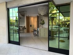 an open glass door leading to a dining room and kitchen area with tables, chairs and chandelier