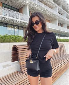 a woman standing next to a wooden bench holding a handbag and wearing black shorts