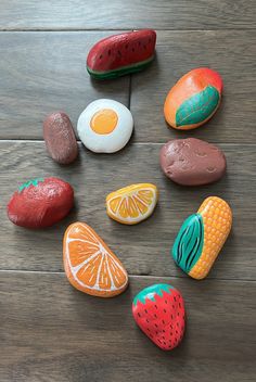 painted rocks arranged in the shape of fruits and vegetables on a wooden surface with an egg, watermelon, orange, strawberry, banana, and other items