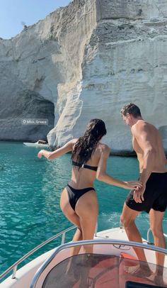 two people in swimsuits standing on the bow of a boat looking at cliffs