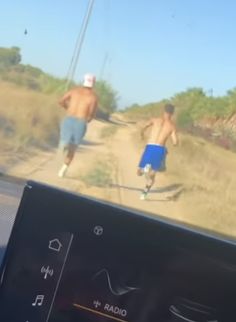 two men running down a dirt road in the sun, one is shirtless and the other is wearing blue shorts