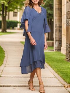 a woman in a blue dress standing on a sidewalk with trees and grass behind her