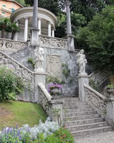 an old building with statues on the steps and flowers in the garden below it, surrounded by greenery