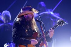 a man with long hair and a hat on singing into a microphone while holding a guitar