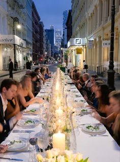 a long table with people sitting at it in the middle of an alleyway, surrounded by tall buildings