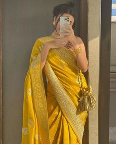 a woman in a yellow sari taking a selfie with her cell phone while wearing gold jewelry