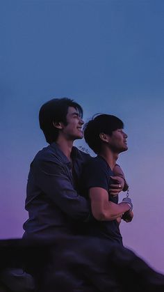 two young men sitting on top of a large rock looking at the sky with their arms around each other