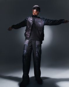 a man standing in front of a gray background wearing black pants and a leather jacket