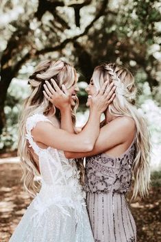 two beautiful women standing next to each other in front of trees and leaves on the ground