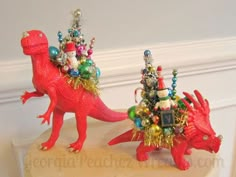 two red dinosaurs with christmas decorations on their heads, one has a tree and the other is a toy dinosaur