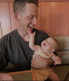 a man holding a baby in his right hand and smiling at the camera while sitting on a couch