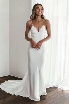 a woman standing in front of a window wearing a white wedding dress with spaghetti straps