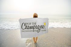 a woman standing on the beach holding a white towel that says pop the champagne i'm changing my name