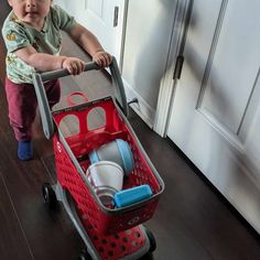 a toddler pushing a red shopping cart with dishes in it's basket on the floor