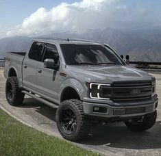 a silver truck parked on top of a cement parking lot next to grass and mountains