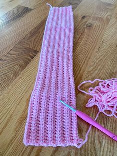 a pink crochet scarf next to a knitting needle on a wooden floor with yarn