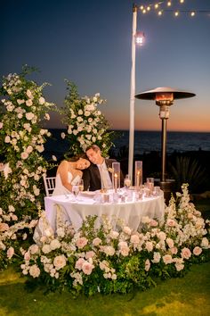 a bride and groom sitting at a table in front of an outdoor setting with flowers