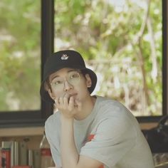 a young man wearing glasses and a hat sitting in front of a window with his hand on his chin