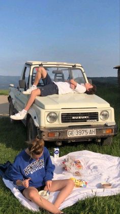 two people laying on a blanket in front of a truck with the hood up and one person sleeping