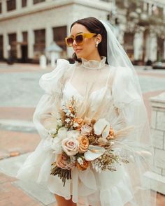 a woman in sunglasses and a white dress is holding a bridal bouquet on the street