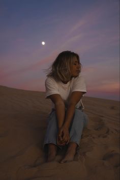 a woman sitting on top of a sandy beach under a purple sky at night with the moon in the distance