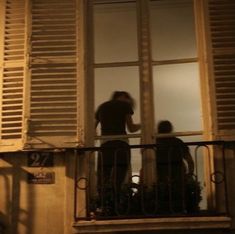 two people standing on a balcony with shutters open and one person looking out the window