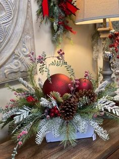 a christmas centerpiece with red balls and greenery on a wooden table next to a lamp