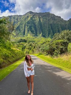 a woman standing on the side of a road in front of a mountain with trees