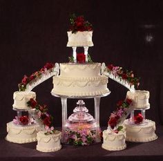 a white wedding cake with red roses on top and tiered cakes below it,