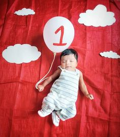 a baby laying on top of a red blanket next to a number one balloon with clouds