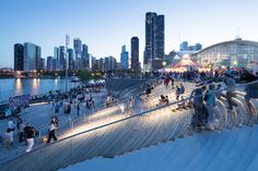 many people are walking around on the boardwalk by the water and buildings in the background