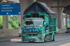 a large green truck driving down a street next to a tall building with a black tarp on it's roof