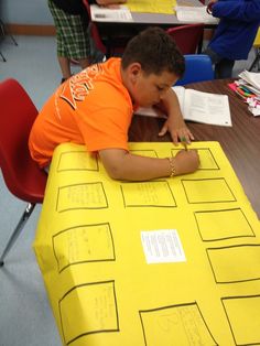 a young boy sitting at a table writing on a piece of yellow paper in front of him