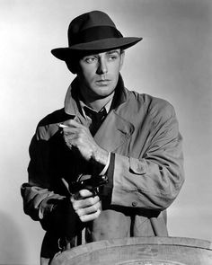 a man in a hat and trench coat is holding a pipe while standing next to a barrel