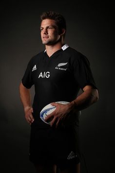 a man holding a rugby ball in his right hand and wearing a black jersey with white letters on it