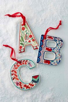 three christmas ornaments are hanging on the snow covered ground, one is decorated with flowers and the other has a letter shaped ornament