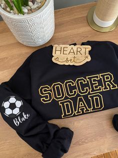 a black sweatshirt with the words soccer dad written on it next to some shoes and a potted plant