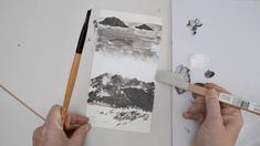 two hands holding a paintbrush next to a painting on a piece of paper with mountains in the background