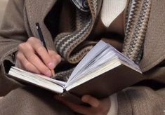 a person sitting down and writing on a book with a pen in their hand while wearing a scarf