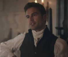 a man wearing a vest and white shirt sitting in a room with candles on the wall