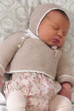 a baby wearing a knitted sweater and bonnet laying on top of a white blanket