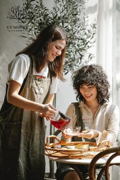 two women sitting at a table pouring something into a glass jar and another woman standing next to her