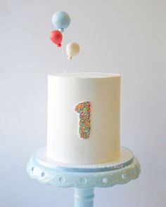 a white cake with sprinkles and balloons on top is sitting on a blue stand