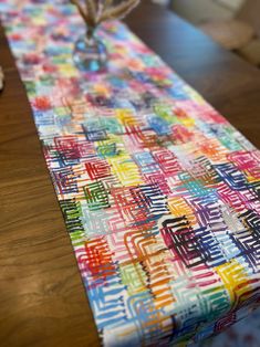 a colorful table runner on top of a wooden table next to a vase filled with flowers