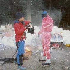 two men in pink snowsuits standing next to a campfire and looking at something on the ground