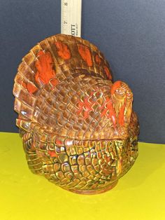 a ceramic turkey sitting on top of a yellow table next to a measuring ruler in the background