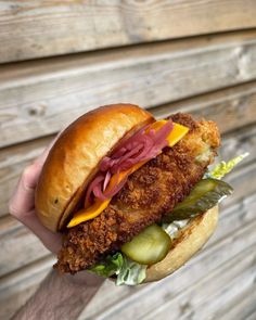 a person holding a chicken sandwich with pickles and cheese on it in front of a wooden wall