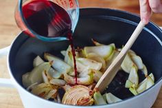 someone pouring red wine into a pot filled with onions and cabbages on a wooden table