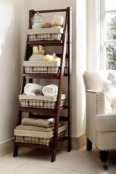 a wooden shelf with baskets and towels on it next to a white chair in front of a window